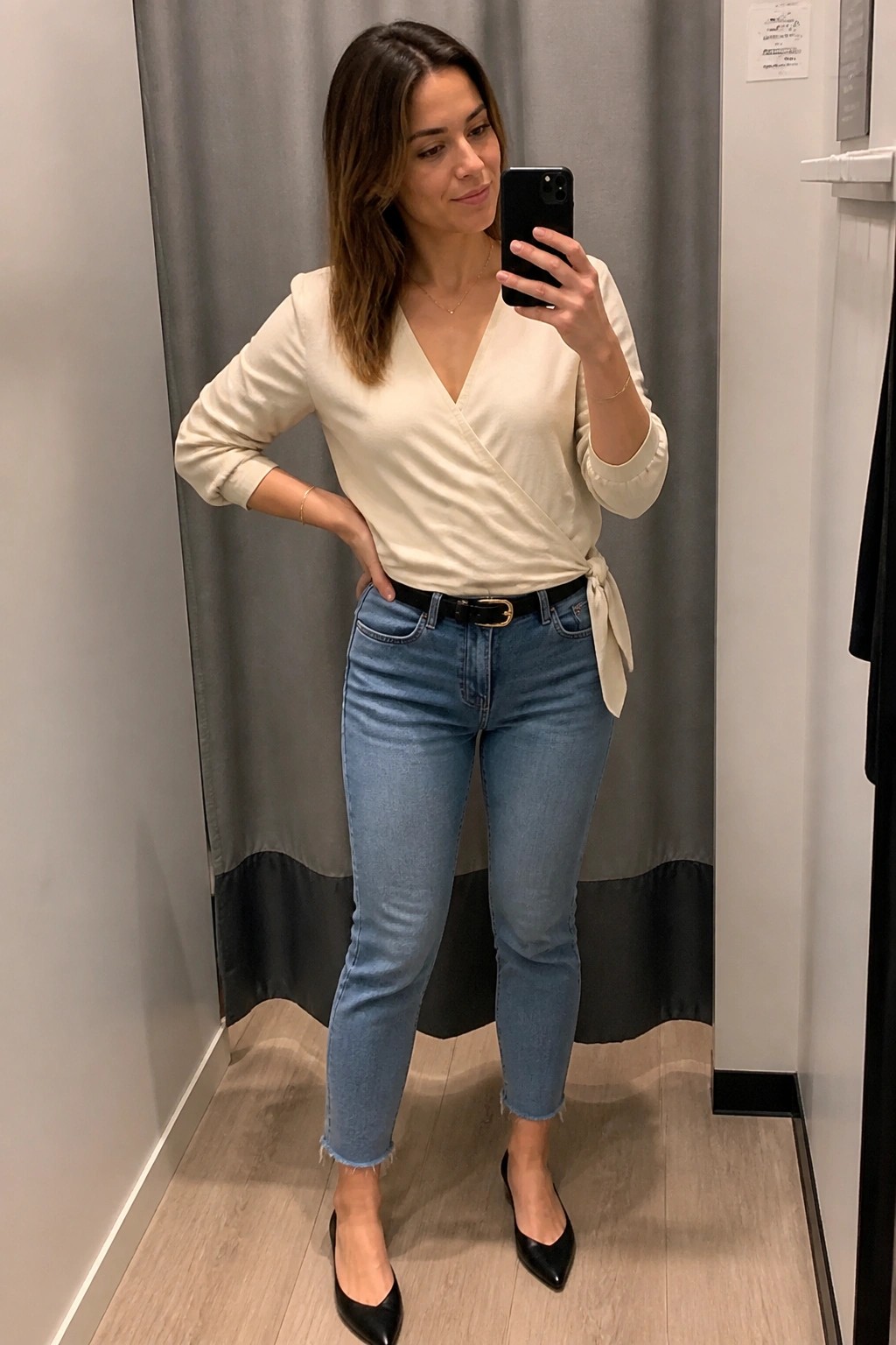 Woman in cream wrap top and blue jeans taking mirror selfie in fitting room