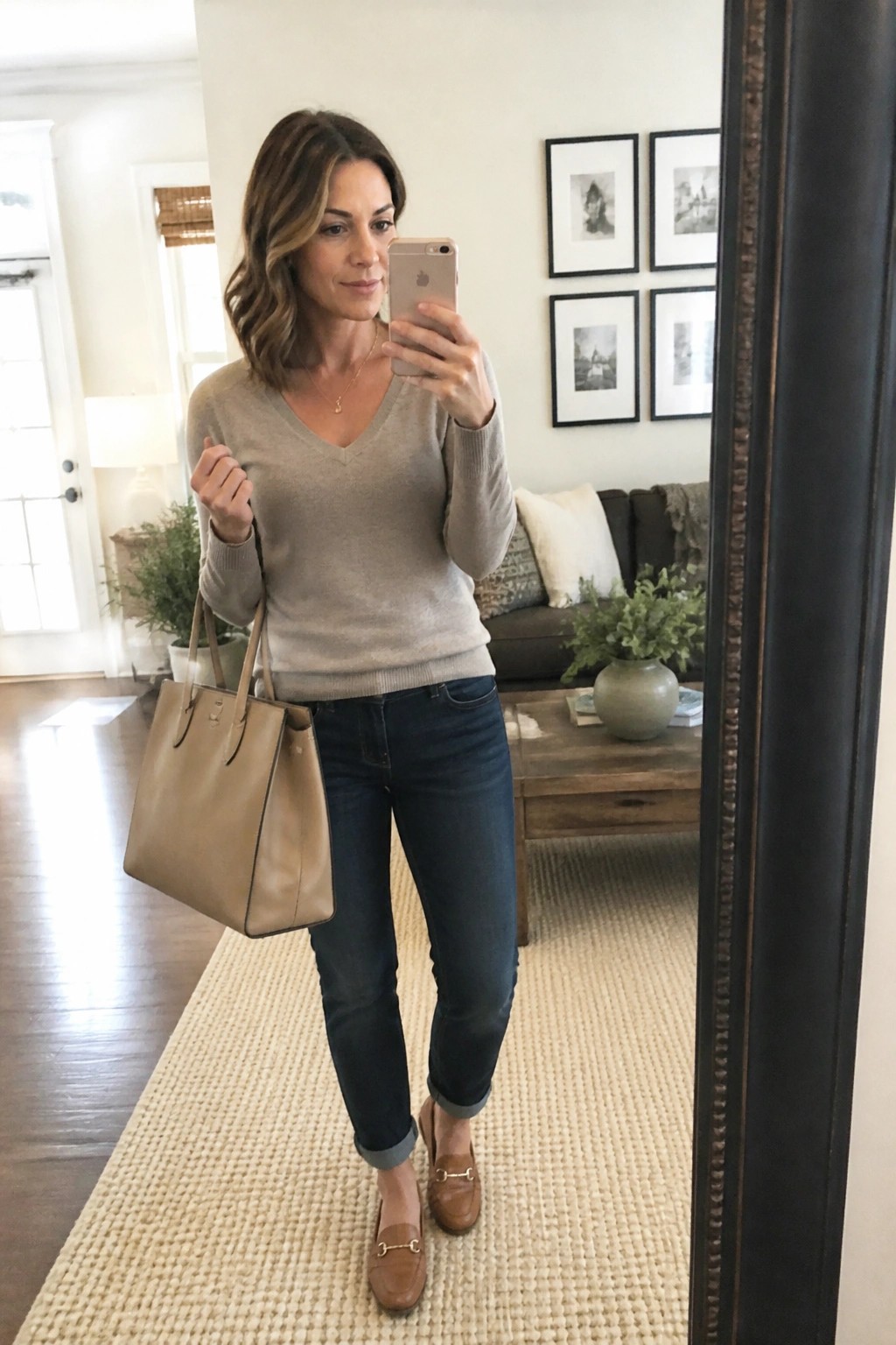 Woman in mirror selfie wearing beige v neck sweater dark blue cuffed jeans brown loafers and carrying matching tote bag while holding phone