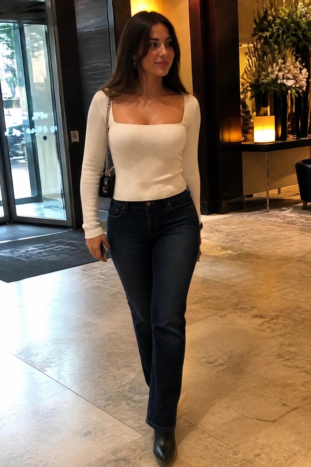 Woman in white square neck ribbed long sleeve top tucked into dark blue straight leg jeans with black pointed boots and small black chain strap handbag.