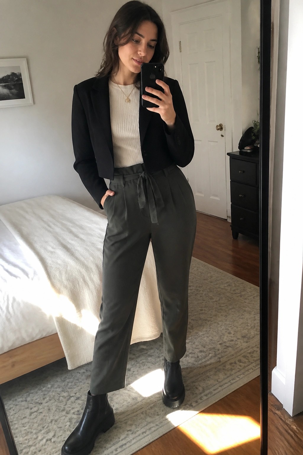 Woman in black cropped blazer and olive pants taking bedroom mirror selfie