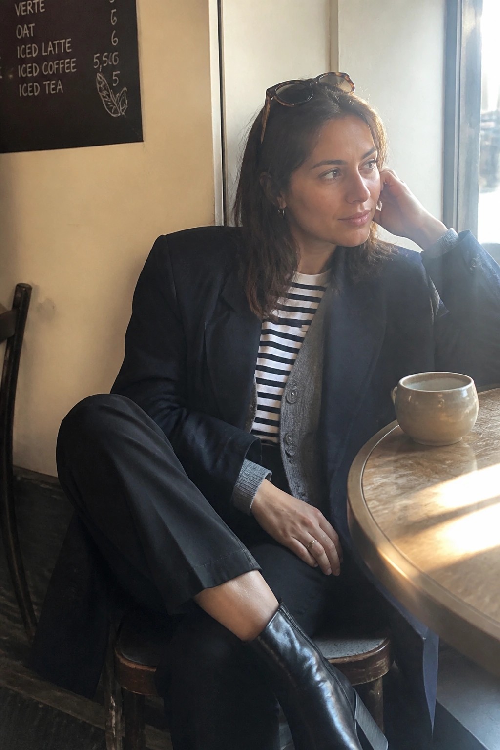 Woman wearing an open black blazer over a black and white striped shirt layered with a gray buttoned vest, paired with black pants and black leather boots.