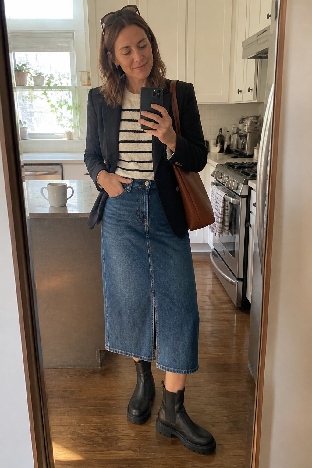Woman in black blazer, striped sweater, denim skirt, and boots taking mirror selfie in kitchen.