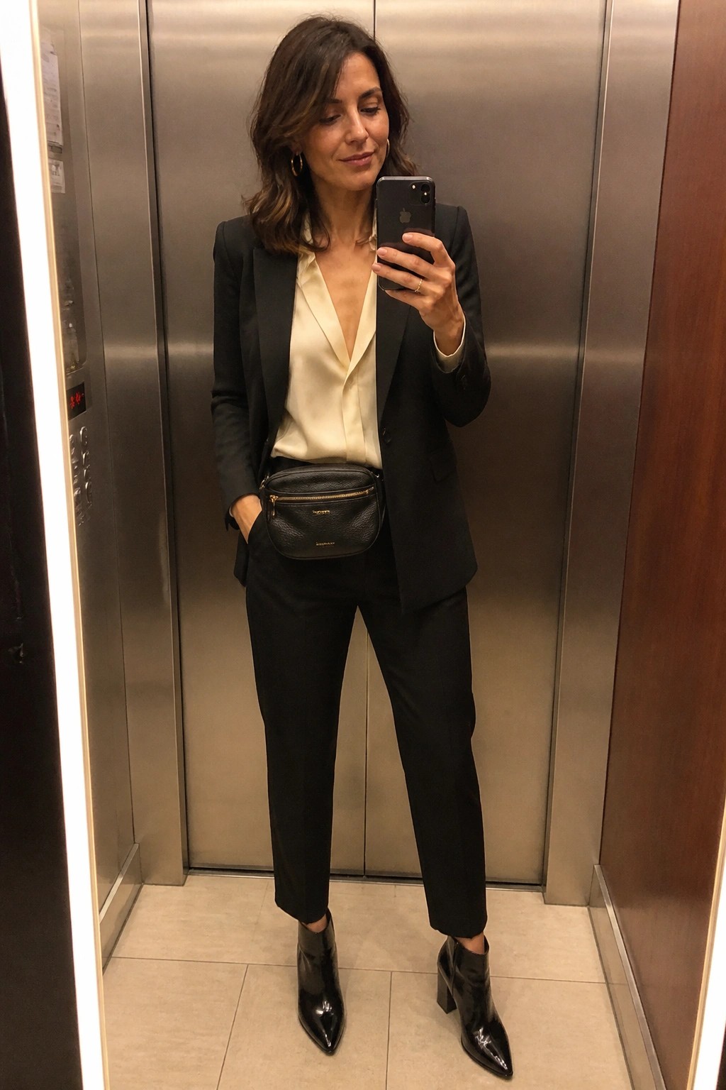 Woman in black suit and cream blouse taking selfie in elevator with belt bag