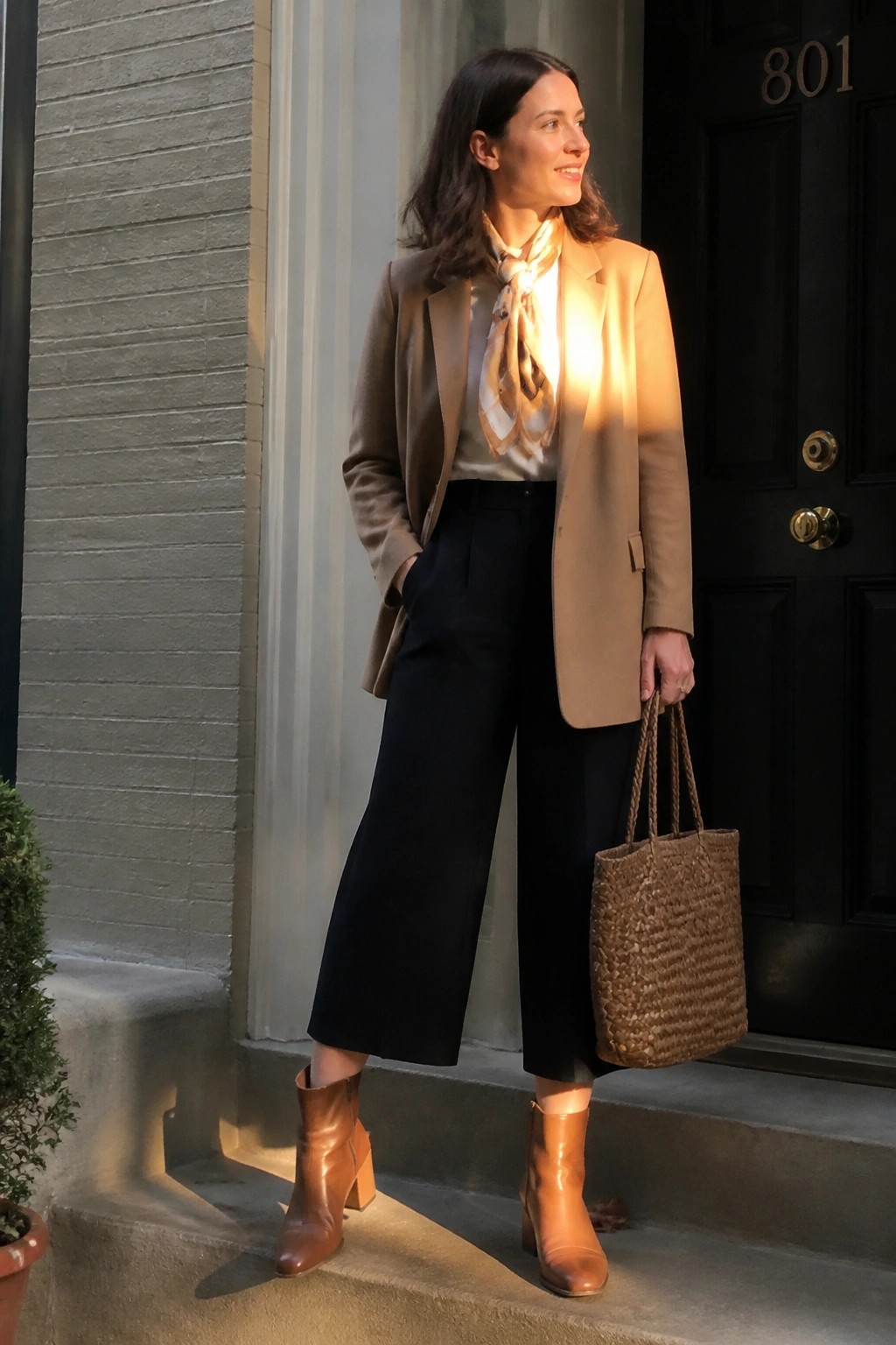 Woman in tan blazer, black culottes, and brown boots holds woven tote on steps