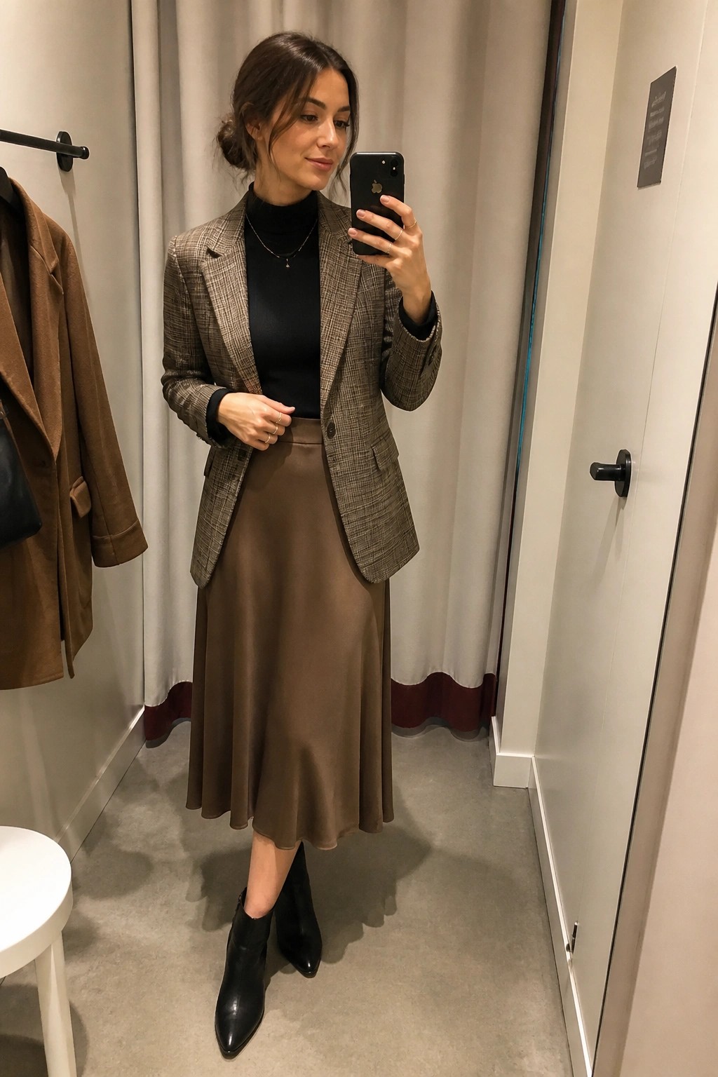 Woman wearing a brown plaid blazer over a black turtleneck, paired with a brown midi skirt and black ankle boots, taking a mirror selfie in a fitting room.