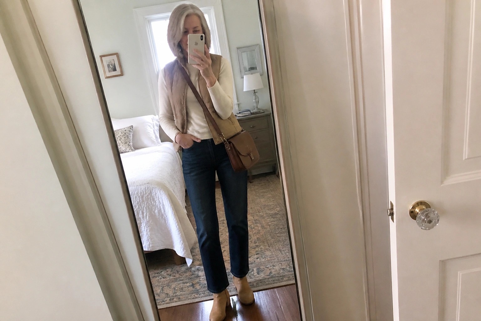 Blonde woman in cream turtleneck and gray puffer vest taking bedroom mirror selfie