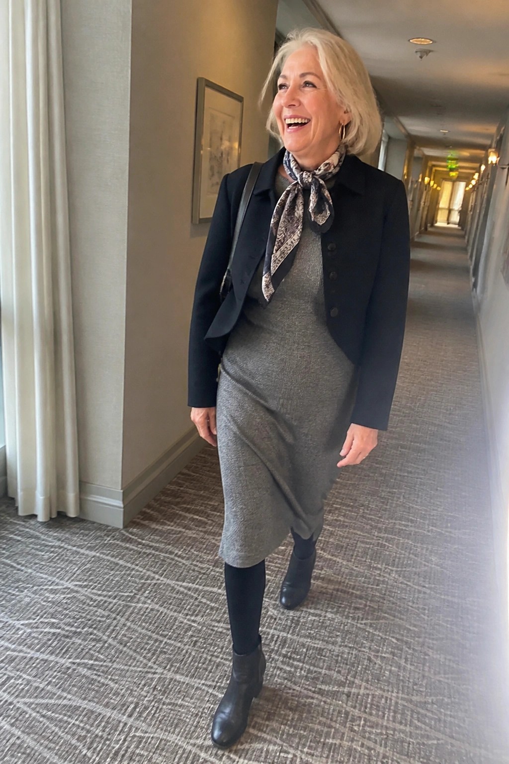 Woman with white hair in black jacket, gray dress, and scarf in hallway