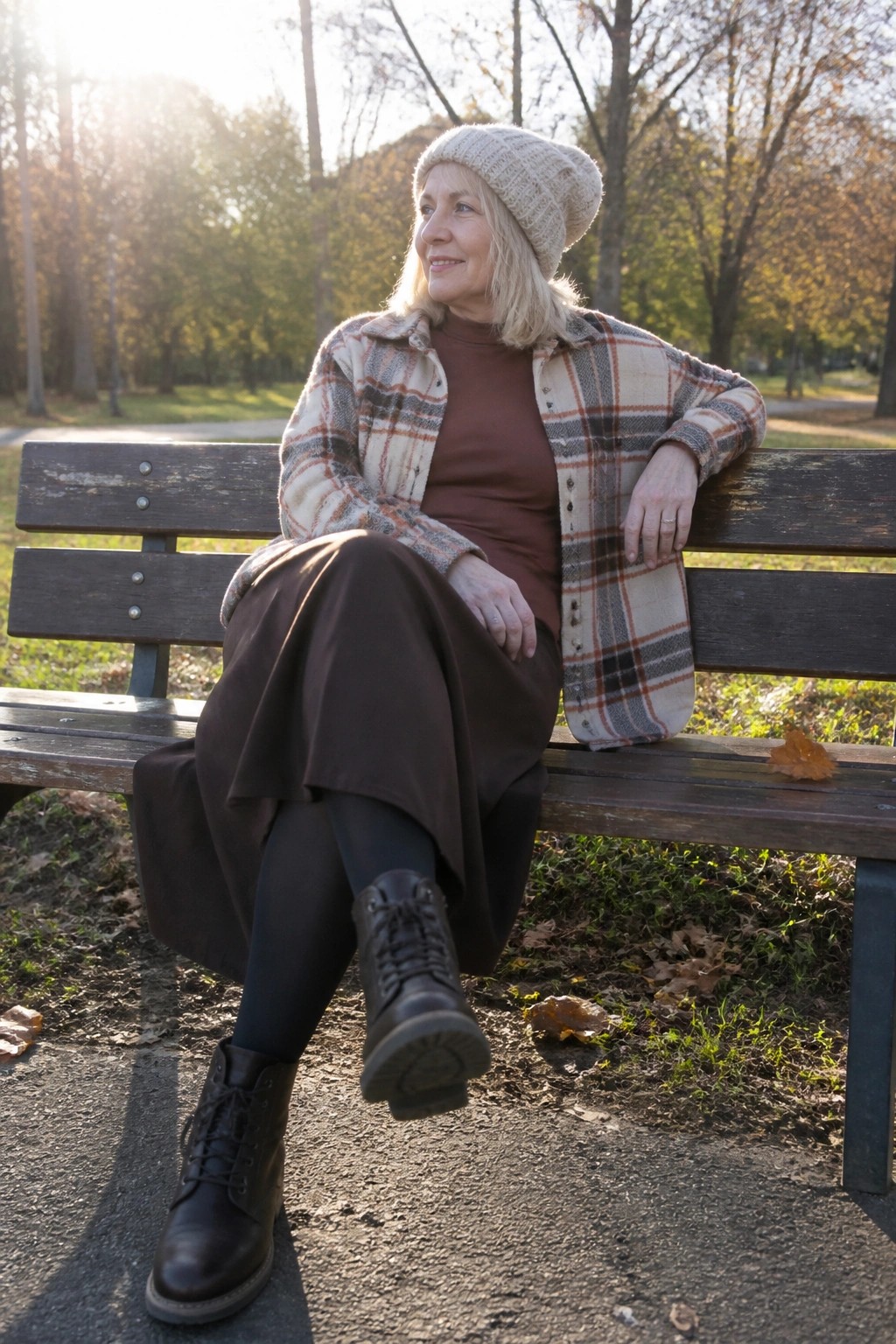 Woman over 60 seated on a bench wearing a beige knit beanie, open plaid button-up shirt in beige brown and black tones over a brown turtleneck, brown midi skirt, black tights, and black lace-up boots.