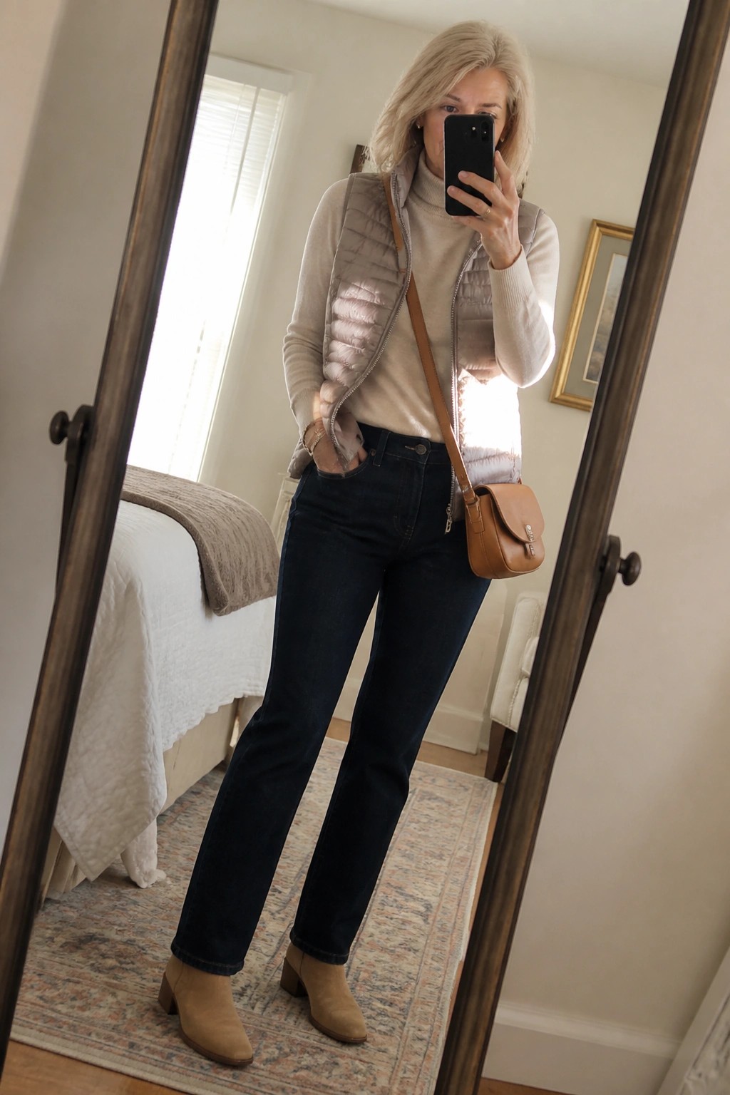 Blonde woman in cream turtleneck and gray puffer vest taking bedroom mirror selfie