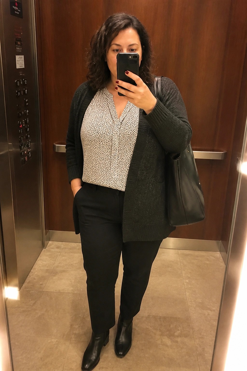A plus size woman wearing a dark cardigan over a patterned blouse with black pants and black boots, taking a mirror selfie in an elevator.