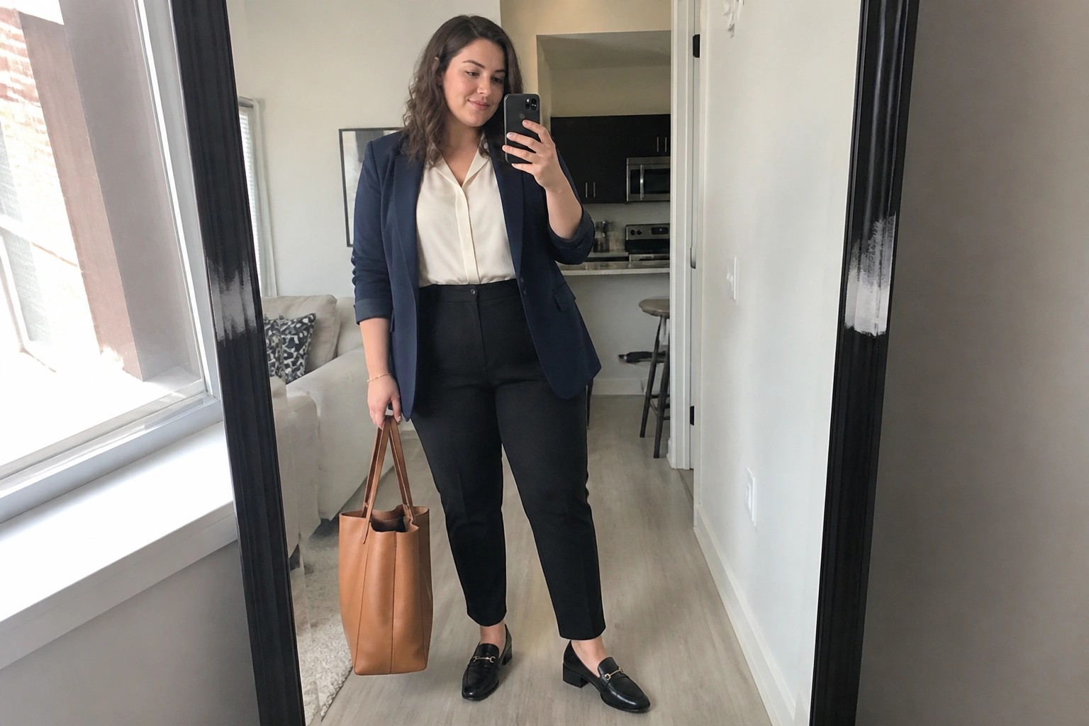 Woman in navy blazer and black pants taking mirror selfie with brown tote bag