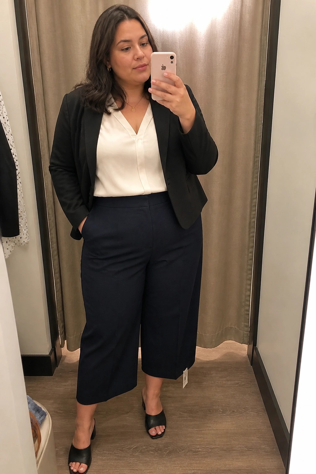 Woman in black blazer and navy pants taking mirror selfie with pink phone