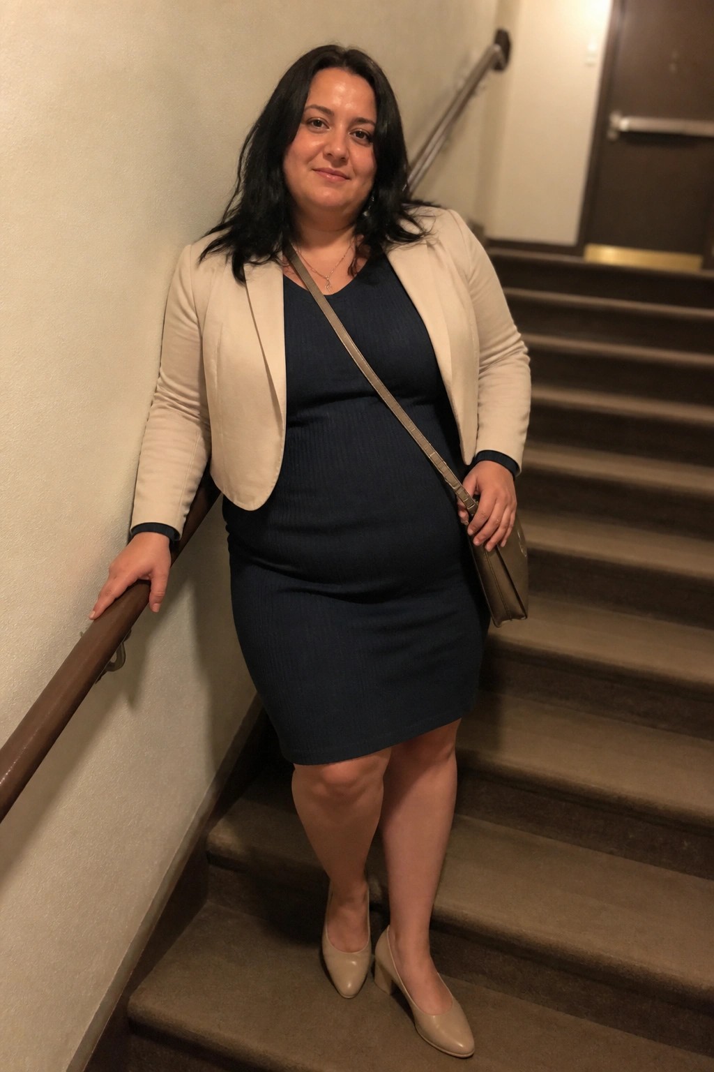 Woman in navy dress and beige blazer standing on indoor staircase with purse