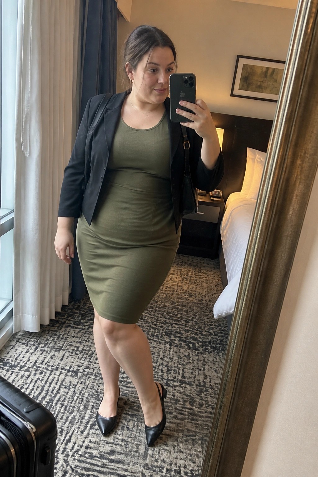 Woman in olive green dress and black blazer taking mirror selfie in hotel room