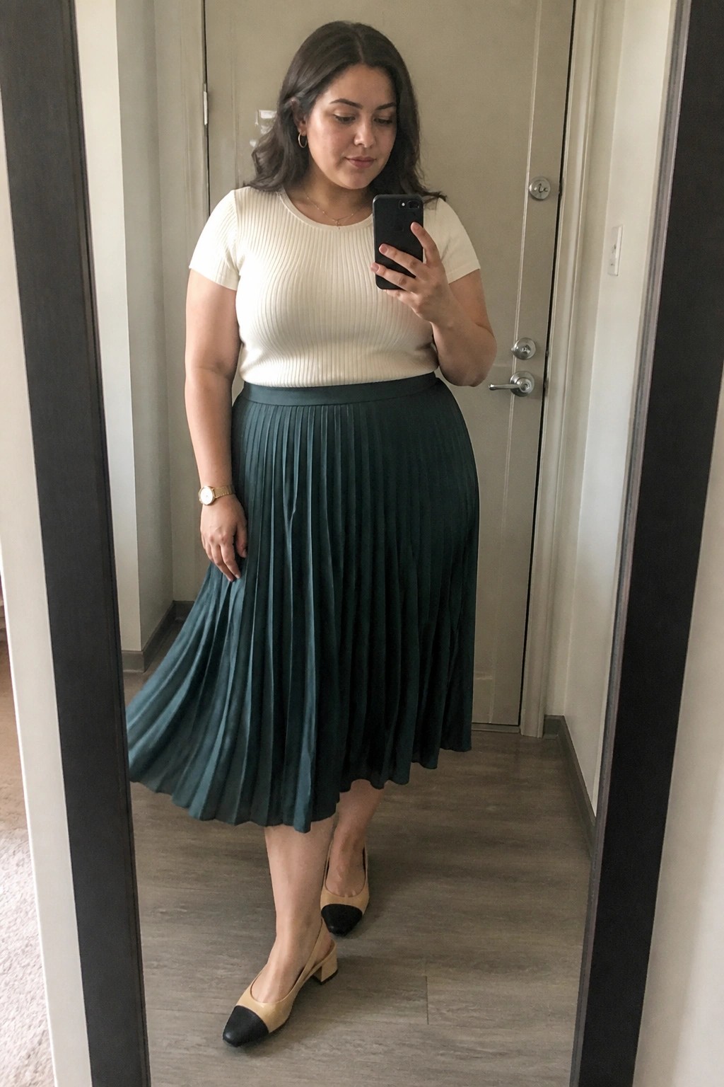 Woman in cream top and teal pleated skirt taking mirror selfie with phone