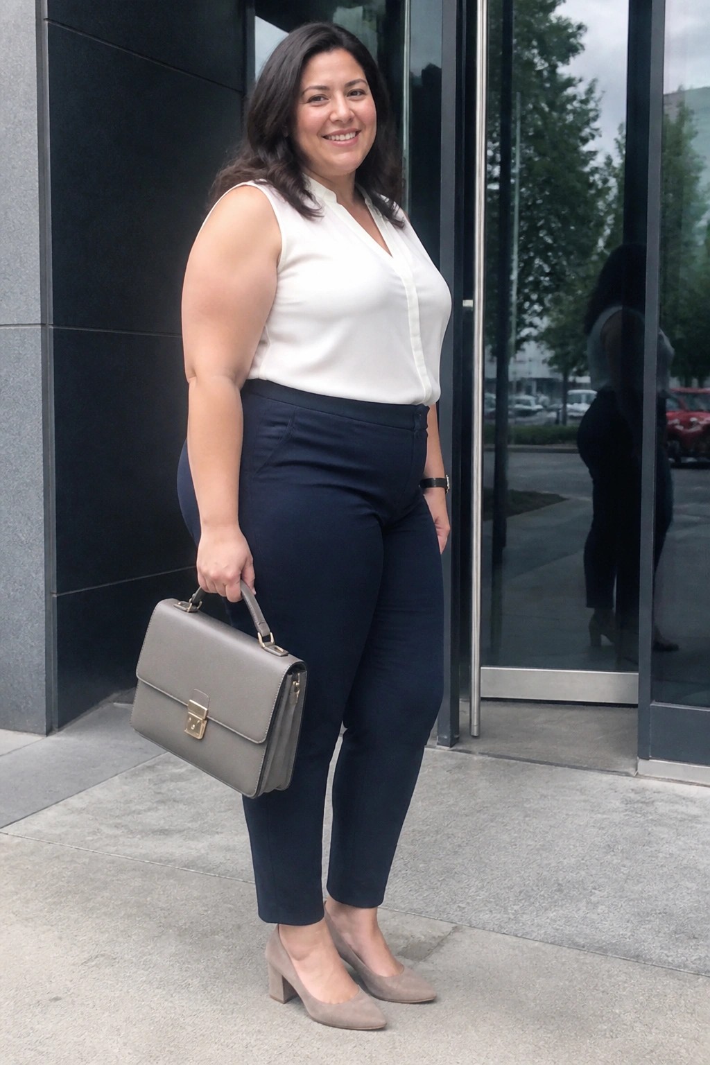 Smiling woman in white blouse and navy pants holds gray handbag outside building.