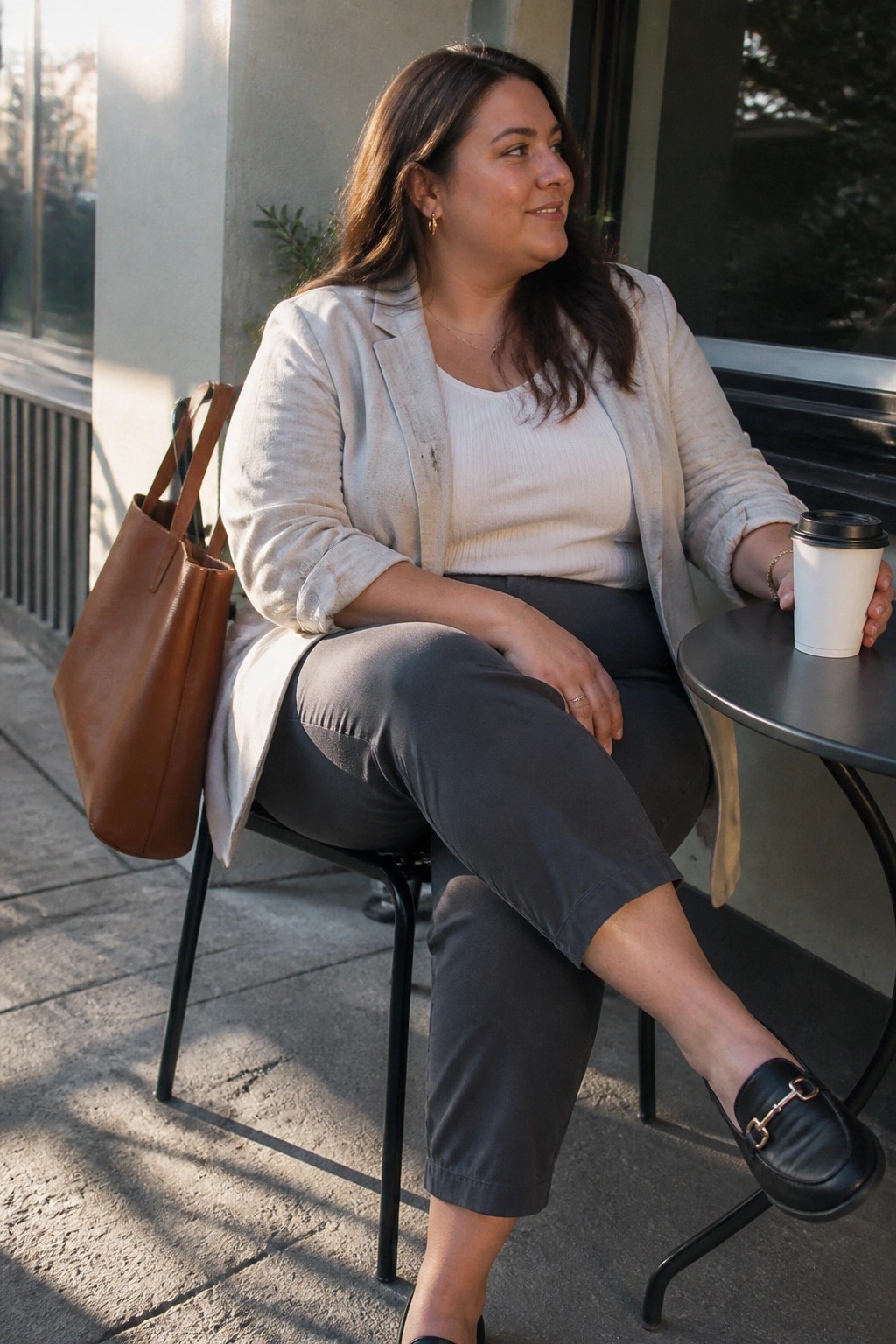 Woman in beige blazer holds coffee at outdoor table with brown leather tote bag.