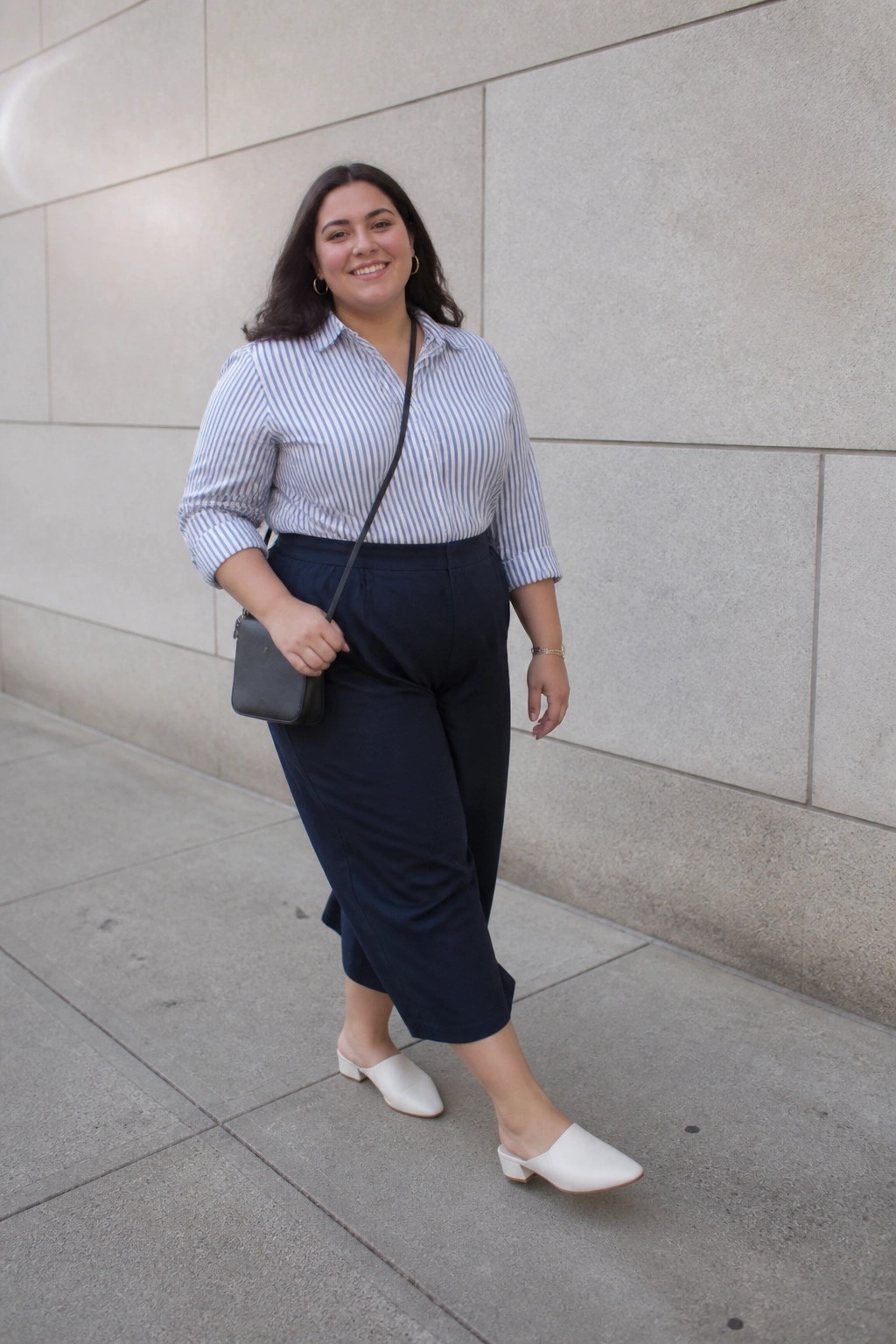 Woman wearing a blue and white striped button-up shirt tucked into navy cropped wide-leg pants with white pointed toe mules and a small black crossbody bag.