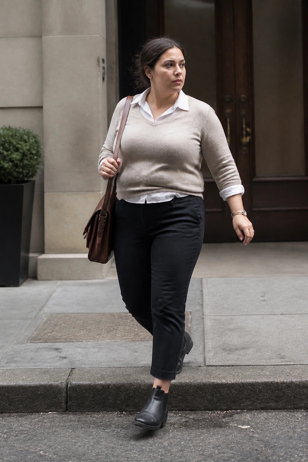 Woman wearing a beige V-neck sweater over a white collared shirt with black pants, black ankle boots, and a brown leather bag.