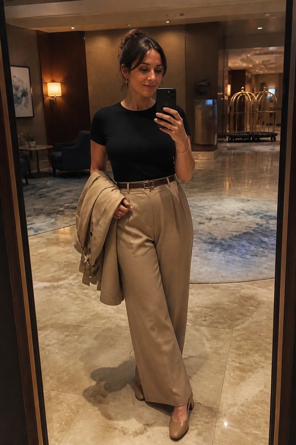 Woman in black short sleeve tee tucked into wide leg taupe trousers with brown belt holding matching blazer over one arm and wearing matching low heels in a hotel lobby mirror selfie.