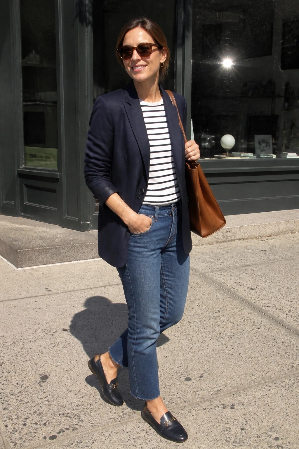 Woman wearing a navy blazer over a black and white striped tee tucked into blue jeans, paired with navy loafers and carrying a brown leather tote bag.