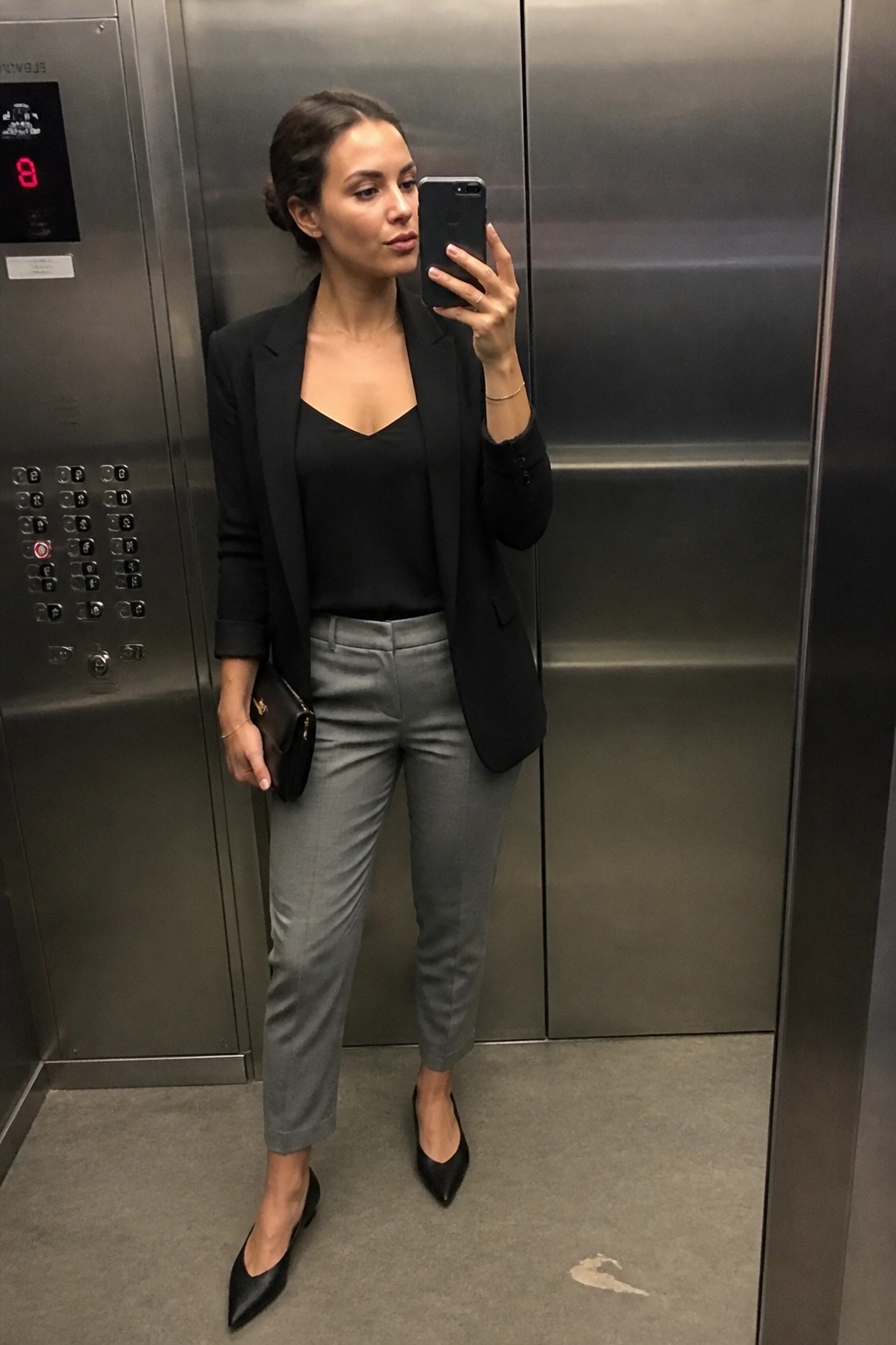 Woman standing in an elevator wearing a black blazer over a black V-neck top with gray trousers and black pointed flats while holding a phone and small clutch.