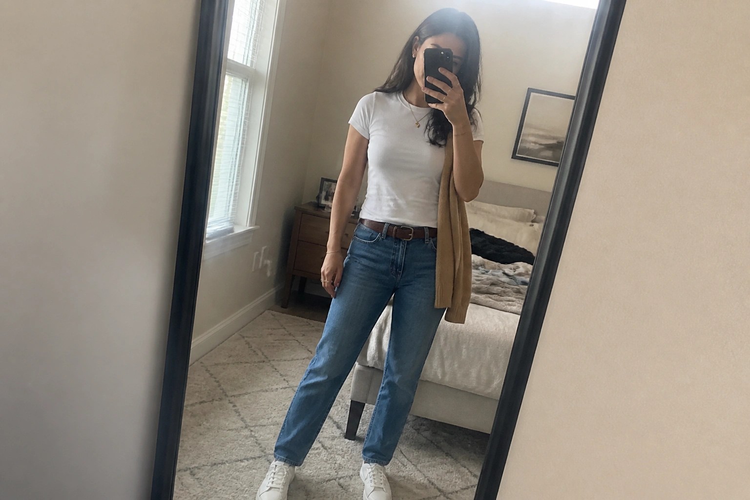Woman in white tee, blue jeans, brown cardigan taking bedroom mirror selfie