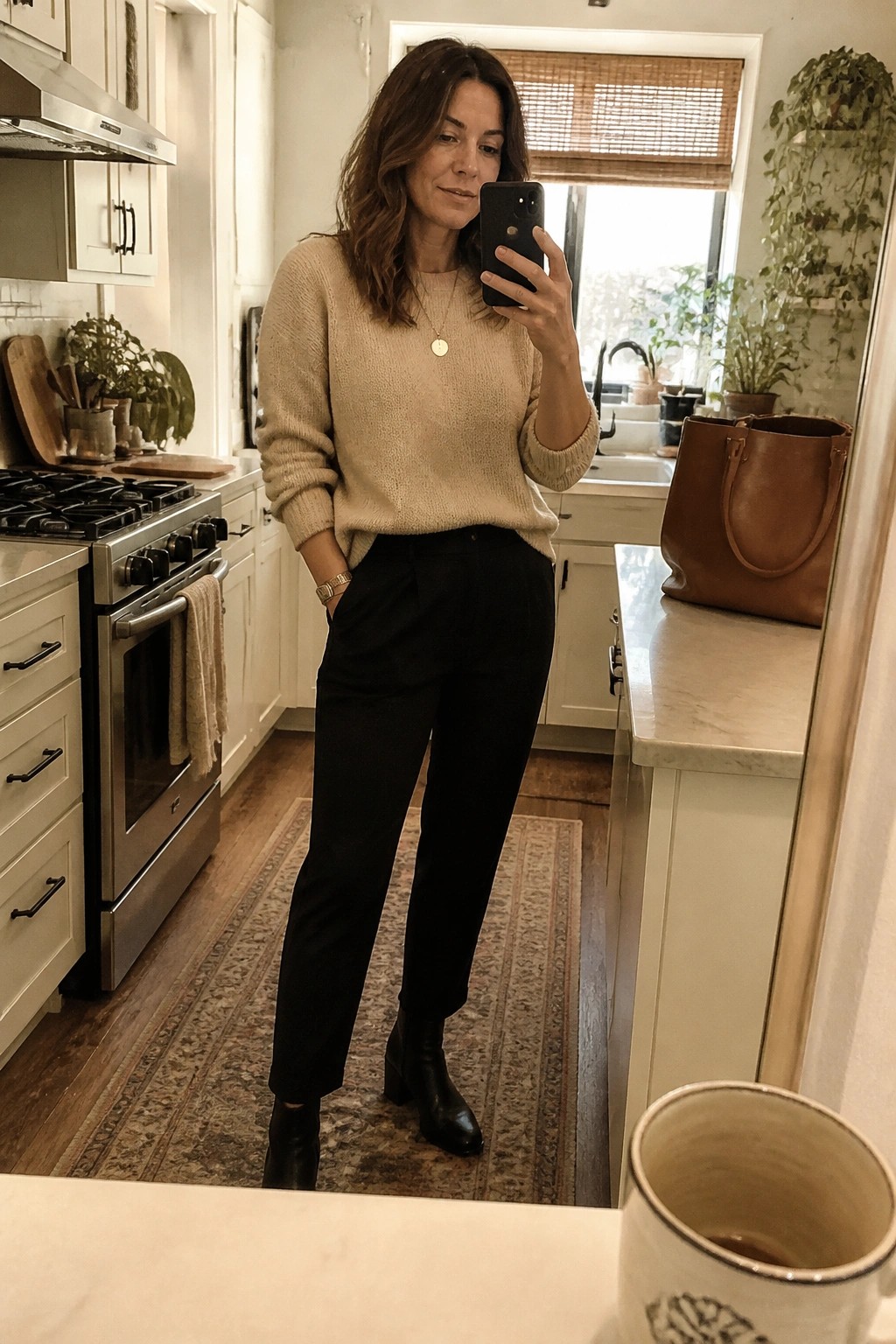 Woman in beige sweater and black pants taking mirror selfie in kitchen