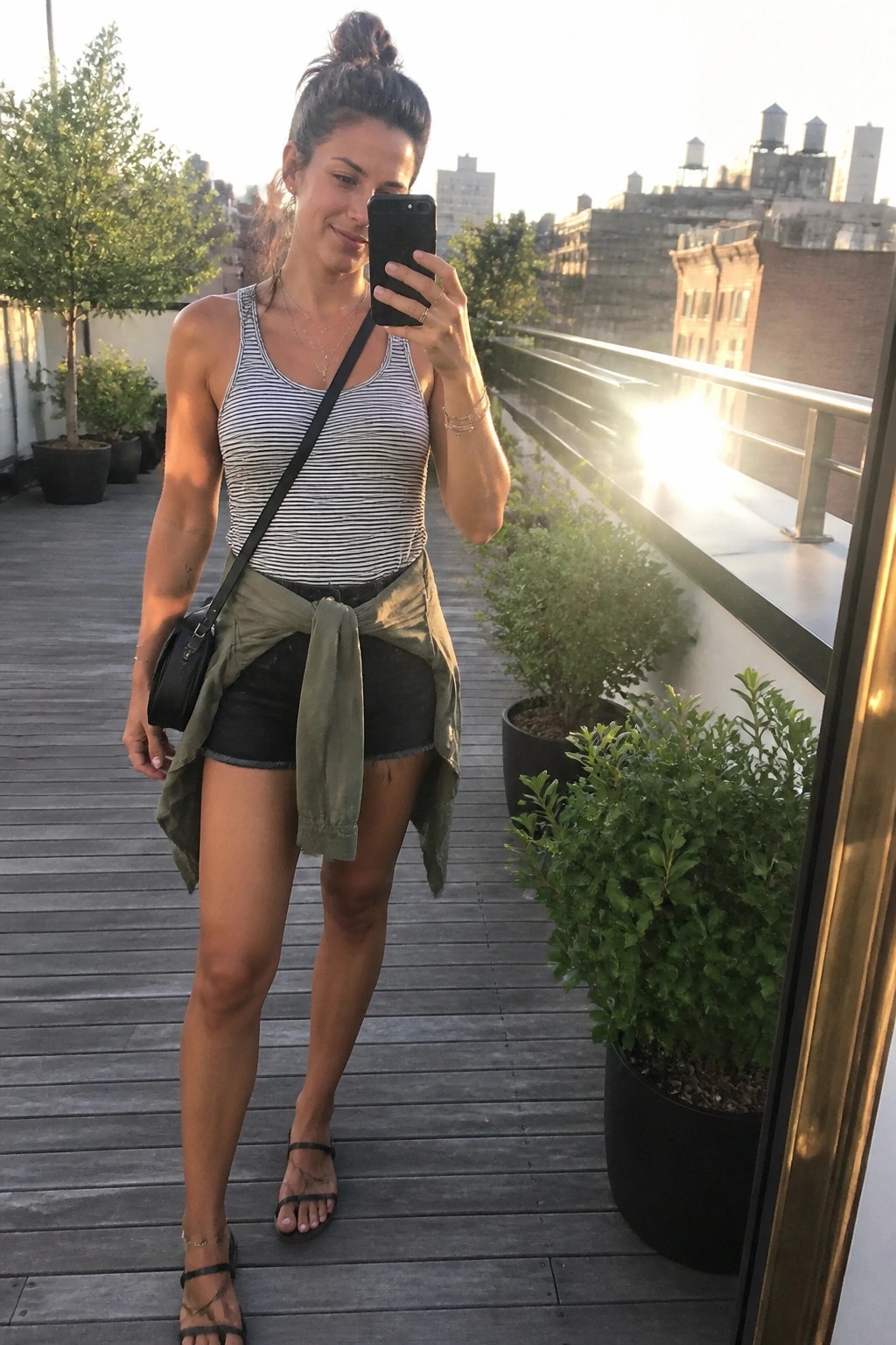 Woman wearing a black and white striped tank top tucked into black denim shorts with an olive green button up shirt tied around her waist, black crossbody bag, strappy black sandals, and layered necklaces on a rooftop deck.
