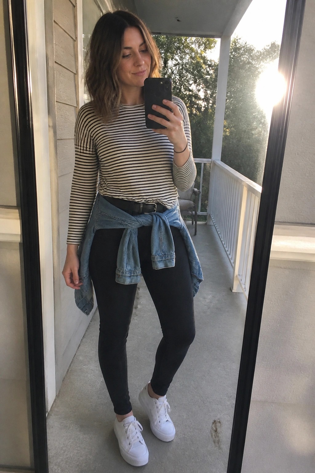 Woman in striped shirt and black leggings with tied denim jacket taking phone selfie on porch