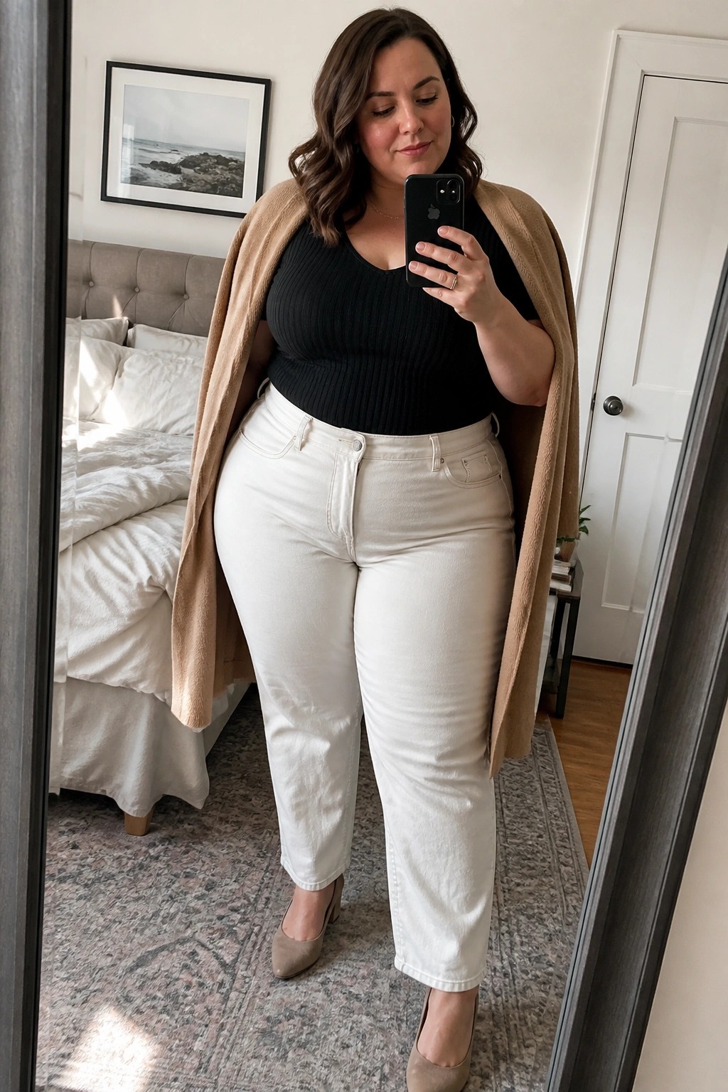 Woman in black top, white jeans, and beige cardigan taking bedroom mirror selfie