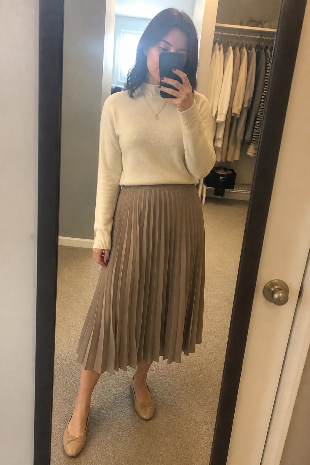 Woman in cream turtleneck and pleated taupe skirt taking mirror selfie with phone
