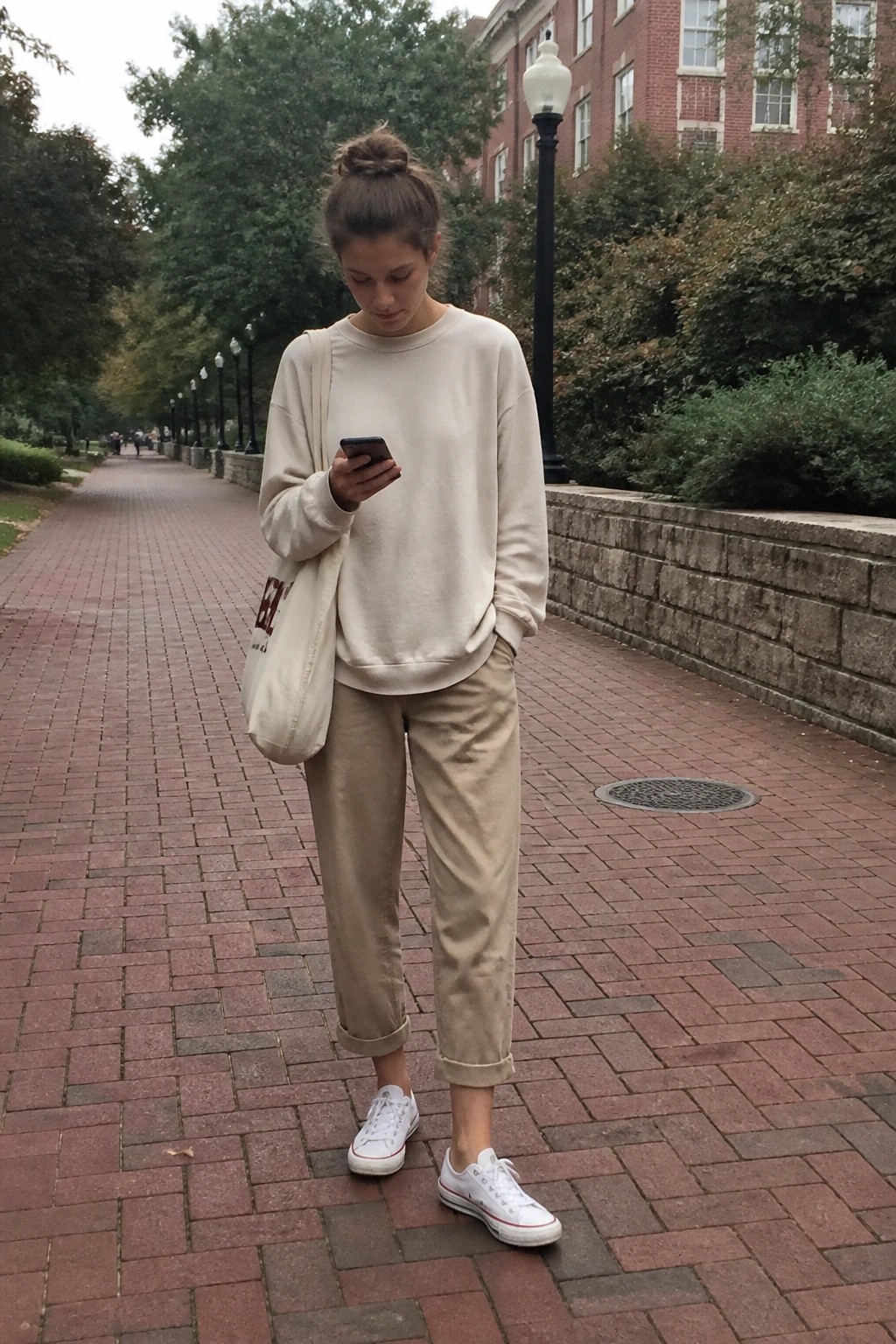 Woman in cream sweatshirt tucked into rolled khaki pants with white low-top sneakers and beige canvas tote bag over one shoulder on a brick walkway.