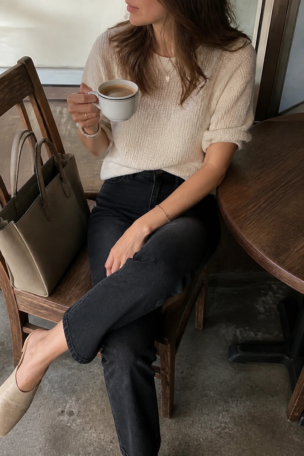 Woman in cream knit sweater sipping coffee beside beige handbag on wooden chair