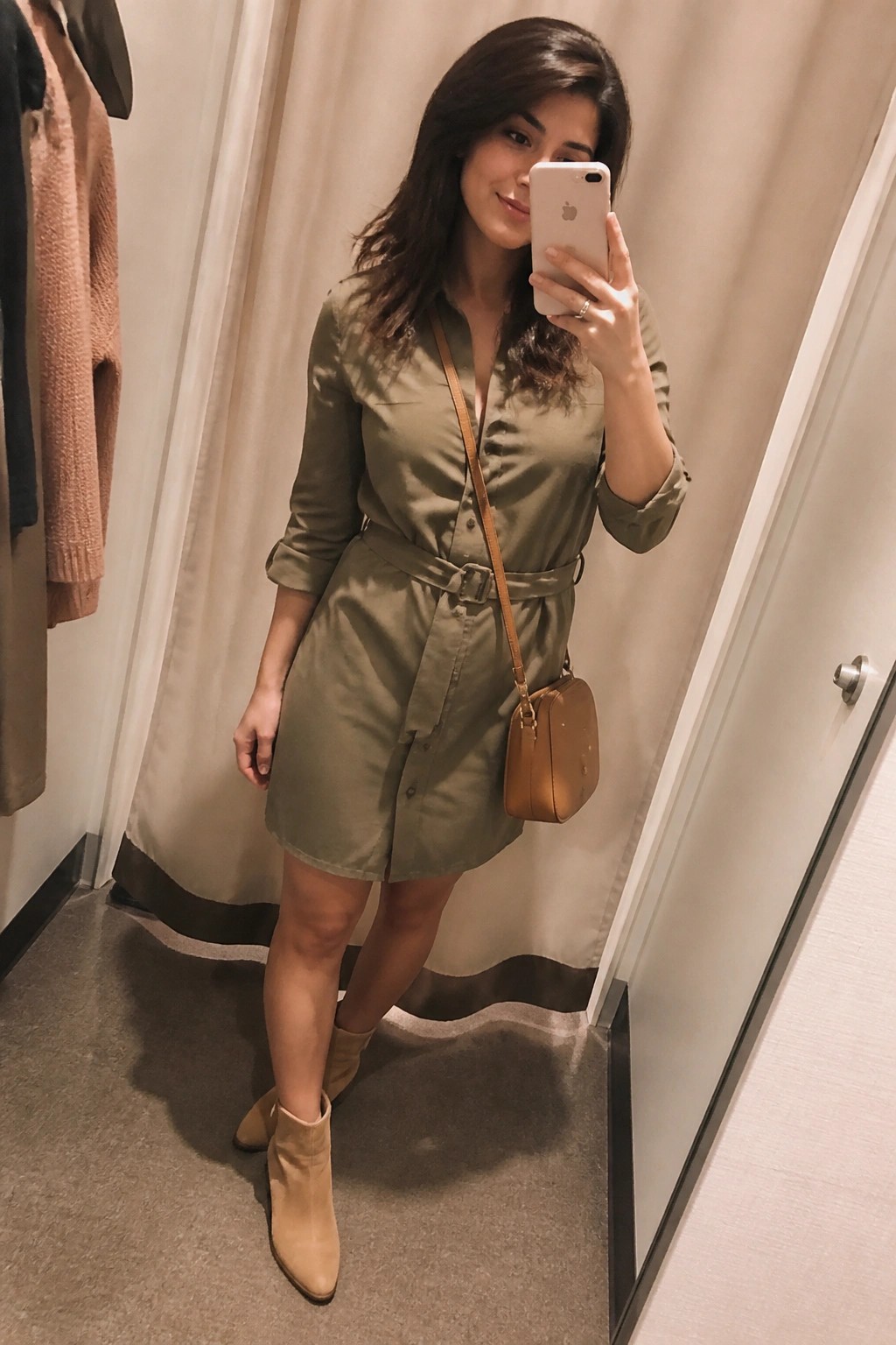 Woman in olive green belted shirt dress and beige boots taking mirror selfie