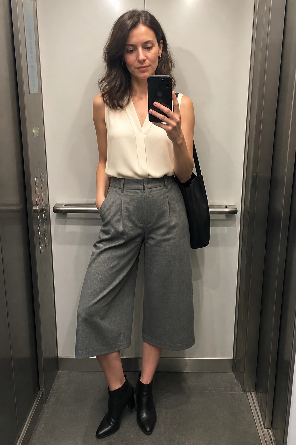 Woman in cream blouse and gray culottes taking selfie in elevator with phone