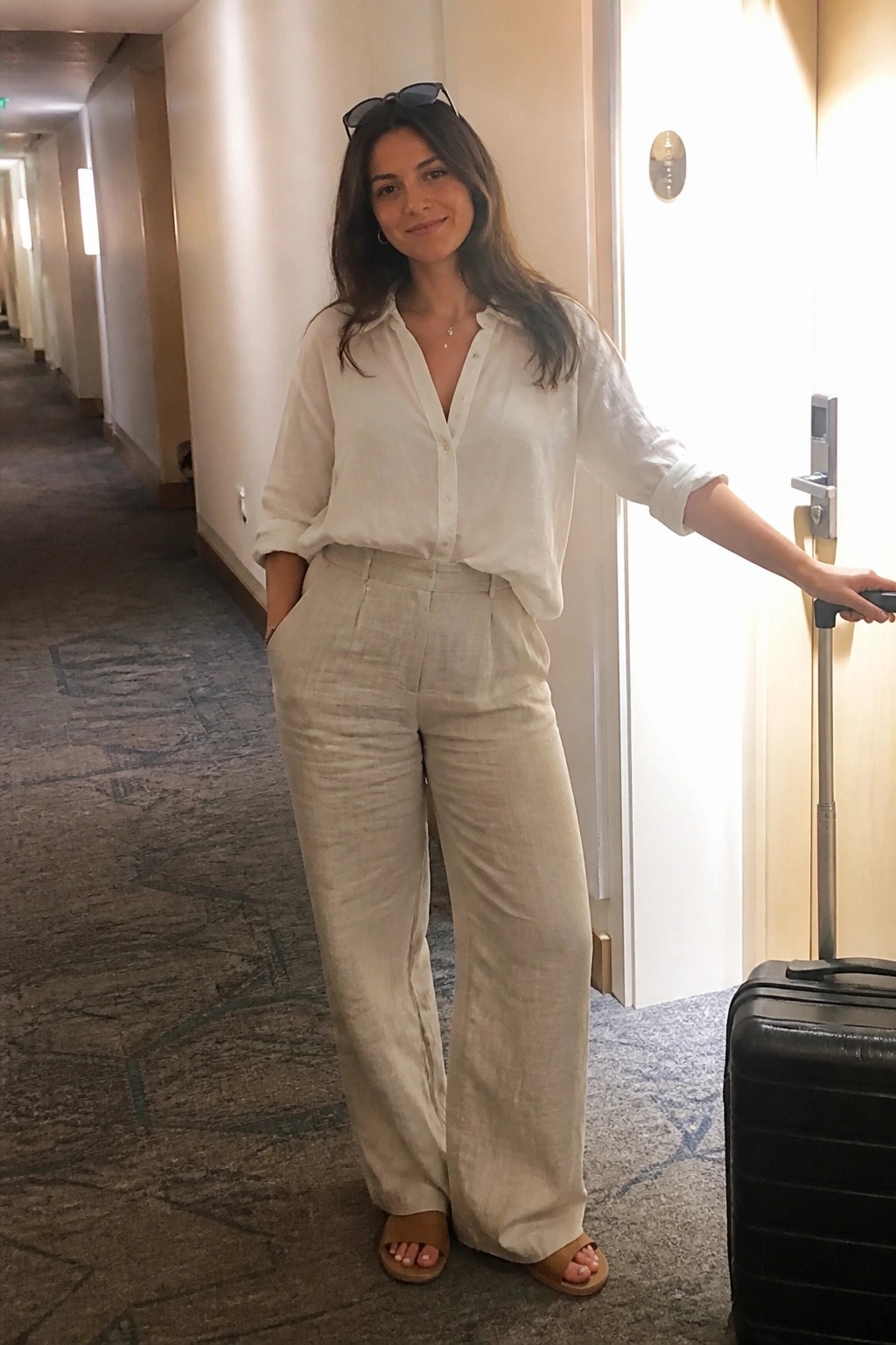 Woman in white shirt and beige pants holding suitcase in hotel hallway