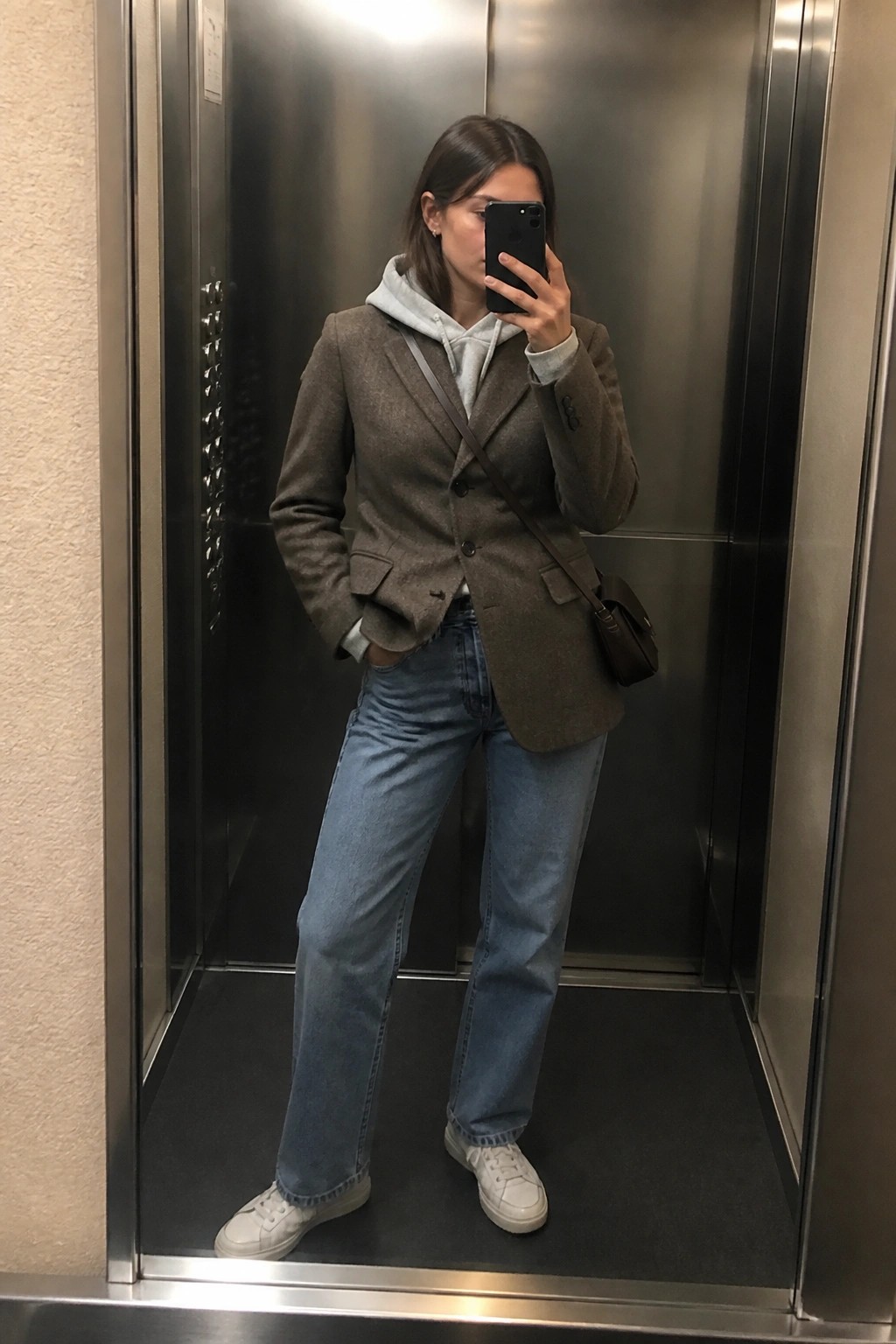 Woman in brown blazer over gray hoodie taking mirror selfie in elevator