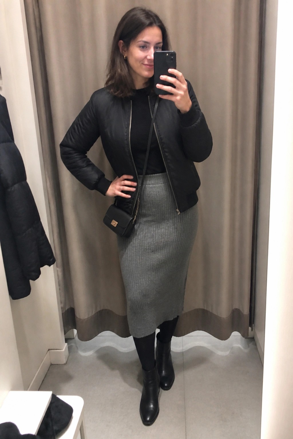 Woman in black bomber jacket and gray midi skirt taking mirror selfie.