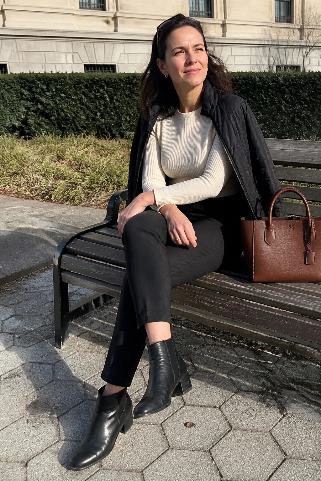 Woman in cream sweater and black jacket on bench beside brown handbag