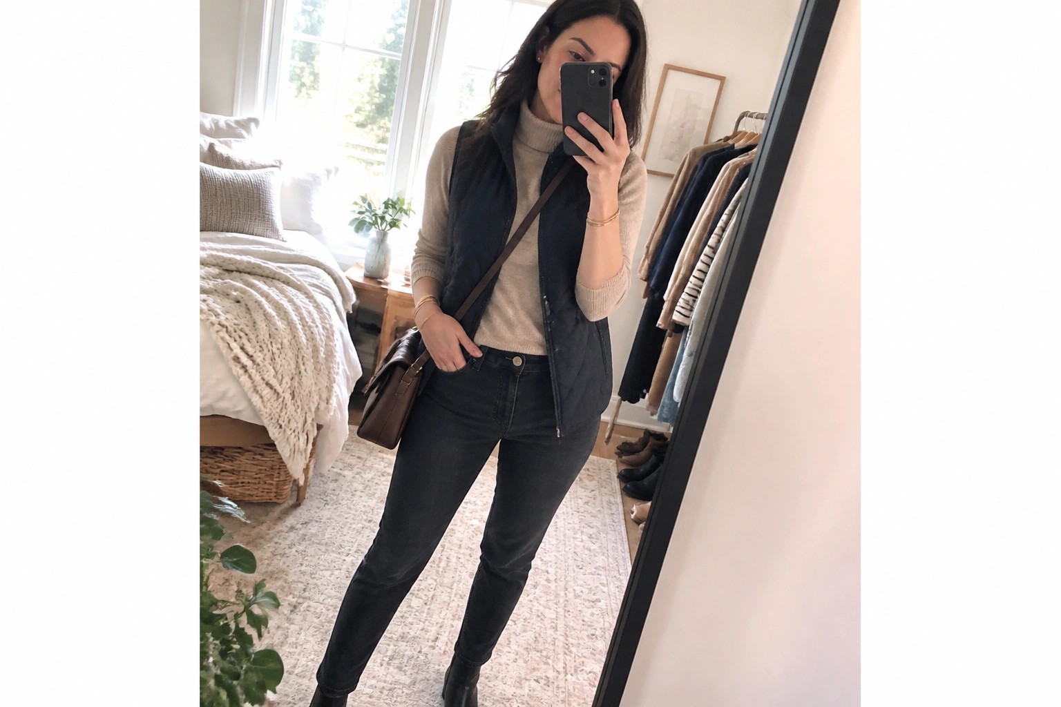 Woman in beige turtleneck, black quilted vest, and blue jeans taking bedroom selfie.