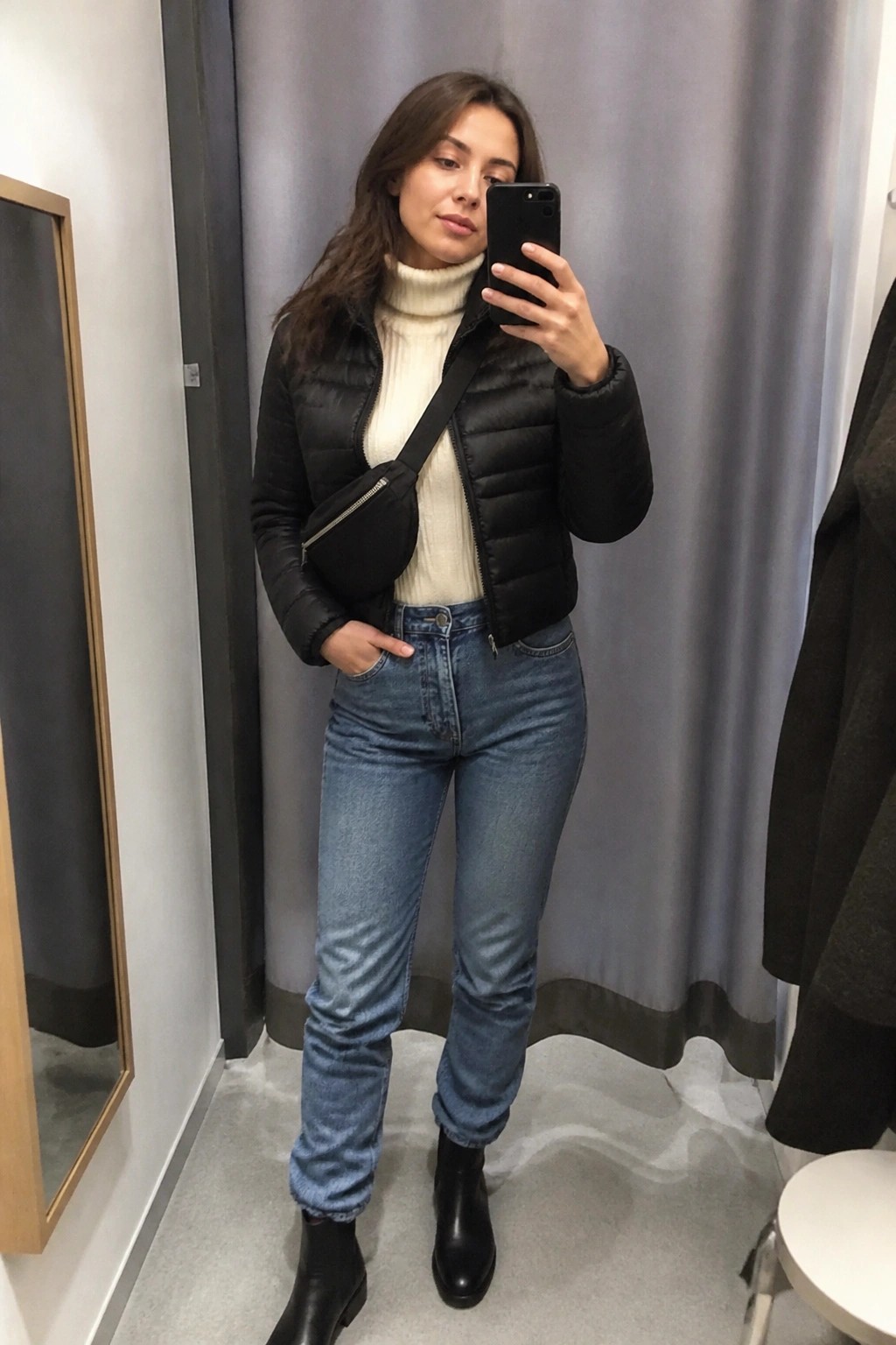 Woman in black puffer jacket and blue jeans taking mirror selfie in fitting room