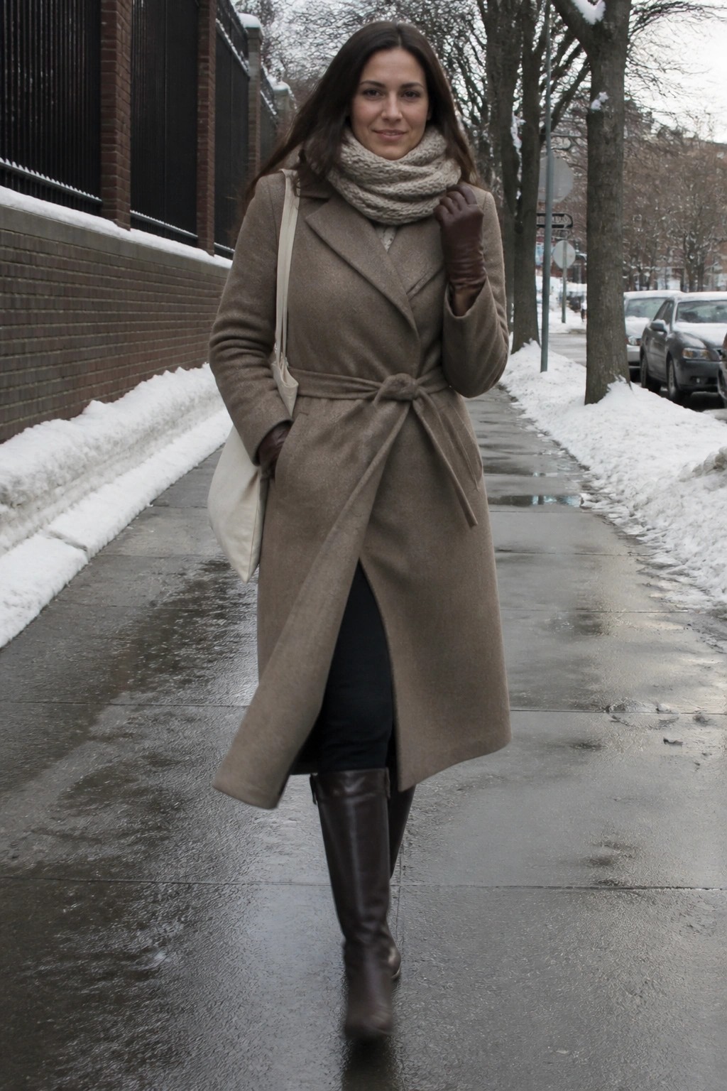 Woman in taupe belted coat, scarf, and boots on snowy sidewalk