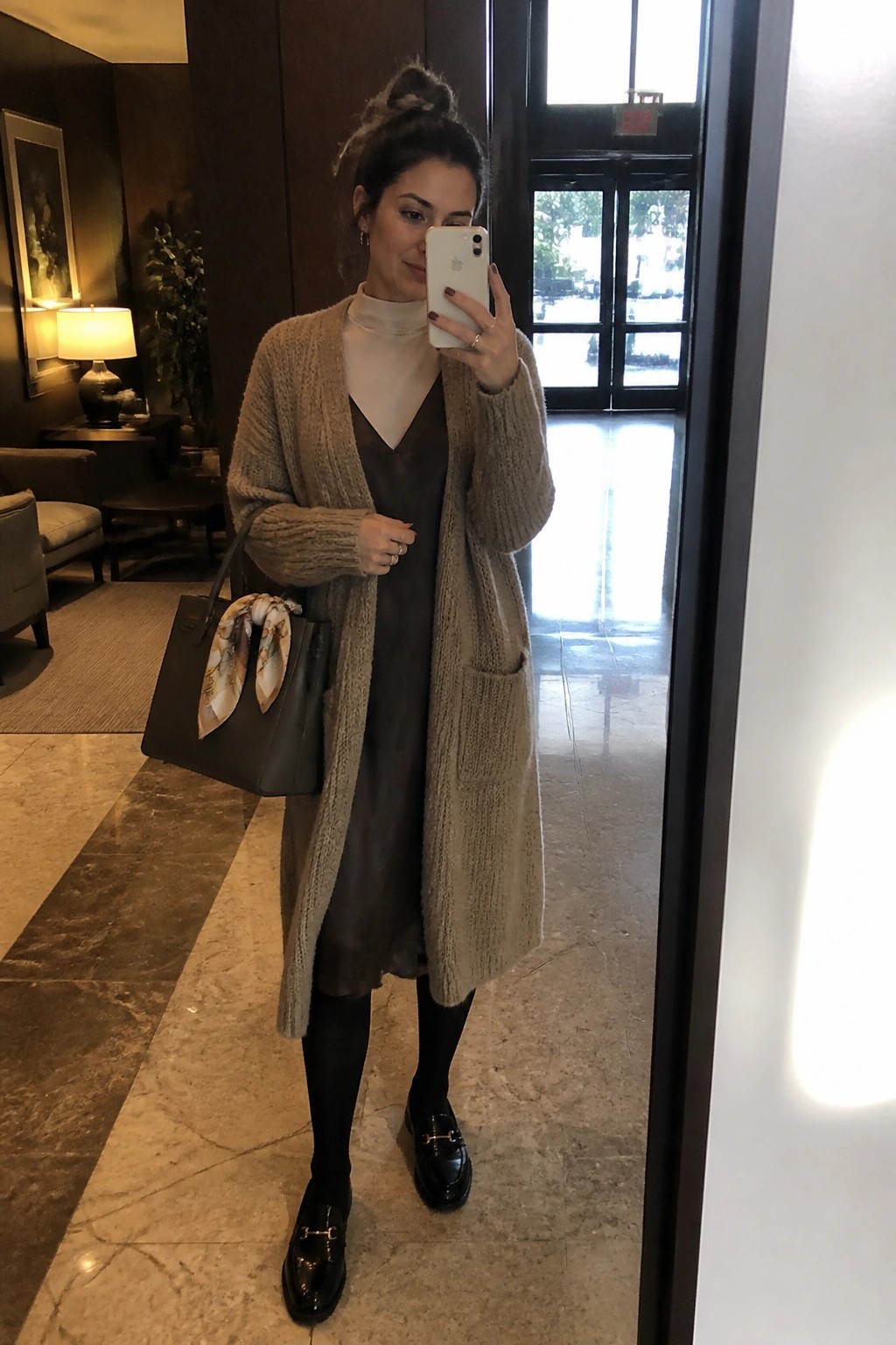 Woman in beige cardigan over brown dress takes mirror selfie in lobby.
