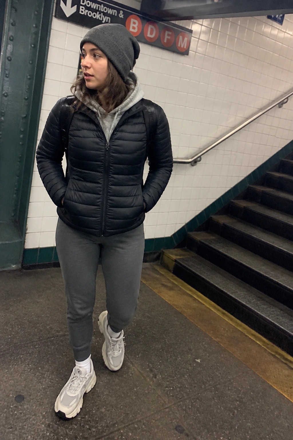 Woman in black puffer jacket and gray beanie stands in subway station near stairs.
