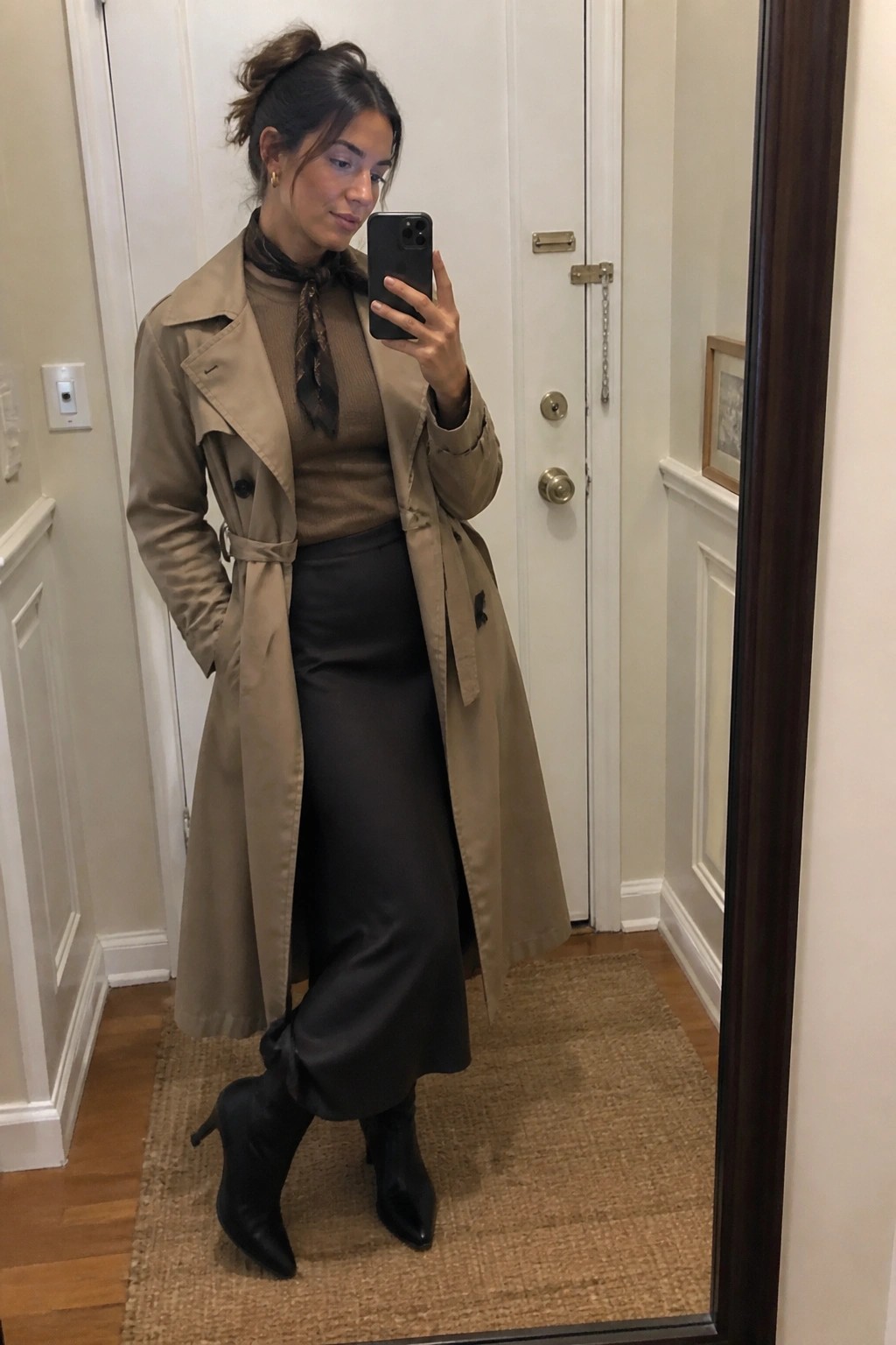Woman in tan trench coat, brown sweater, black skirt, and boots taking mirror selfie