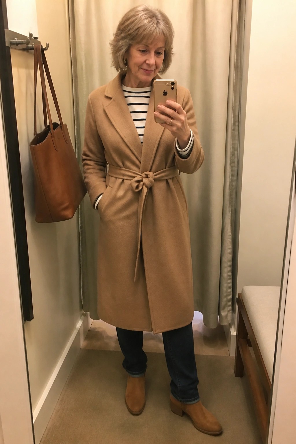 Woman wearing a belted camel wool coat over a navy striped top with dark jeans and tan suede ankle boots while holding a phone in a fitting room with a brown leather tote visible nearby