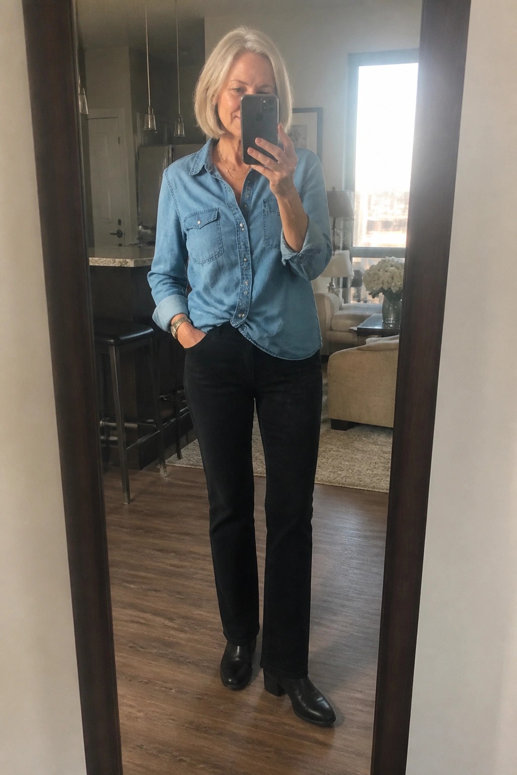 Woman over 60 wearing a light blue denim shirt tucked into black straight leg pants with black heeled ankle boots and a thin gold necklace while taking a mirror selfie.
