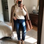 Blonde woman in beige trench coat and jeans taking bedroom mirror selfie with tote bag.
