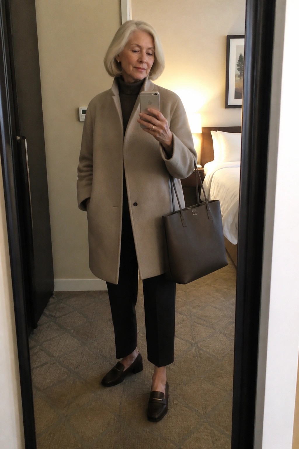 Elderly woman in beige coat taking mirror selfie with brown handbag and phone